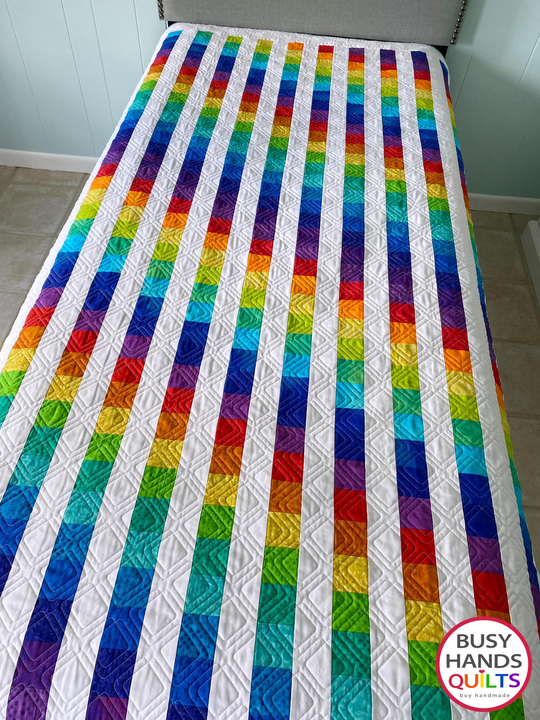 Colorful rainbow twin bed quilt with 'Busy Hands Quilts' branding on a bed.