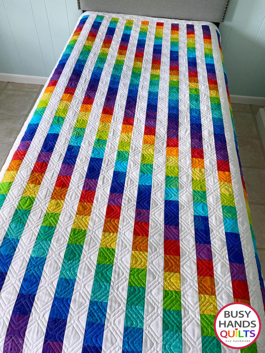 Colorful rainbow twin bed quilt with 'Busy Hands Quilts' branding on a bed.