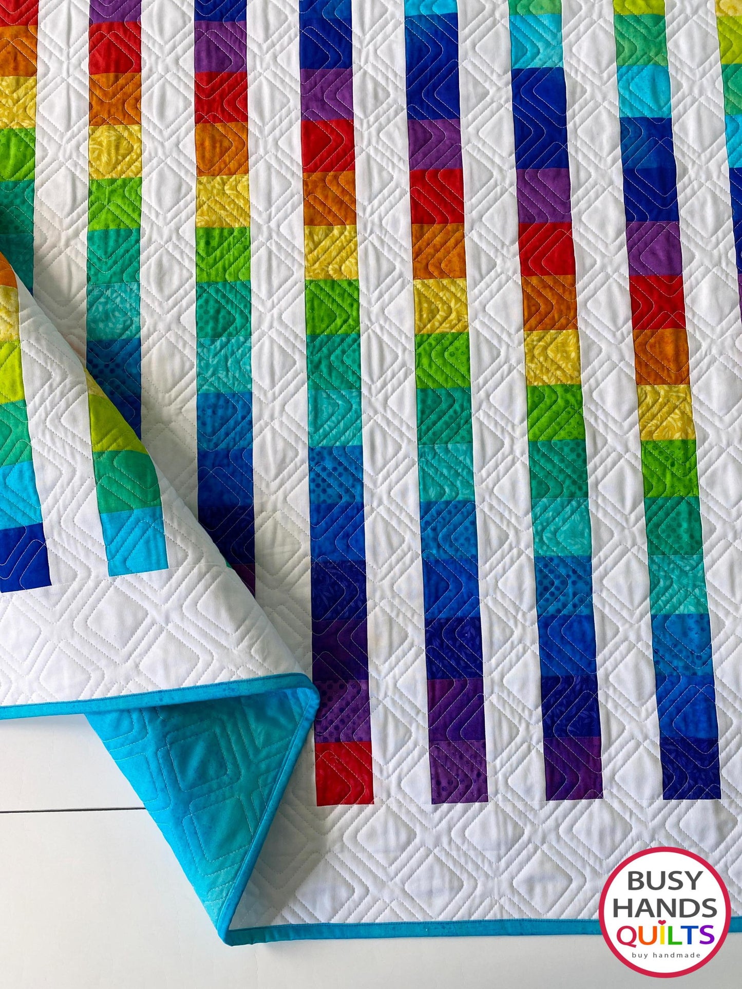 Rainbow quilt with geometric pattern on a white background, featuring the 'Busy Hands Quilts' brand.